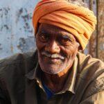 A senior Indian man wearing an orange turban smiles warmly while seated outdoors in Cumbum, India.